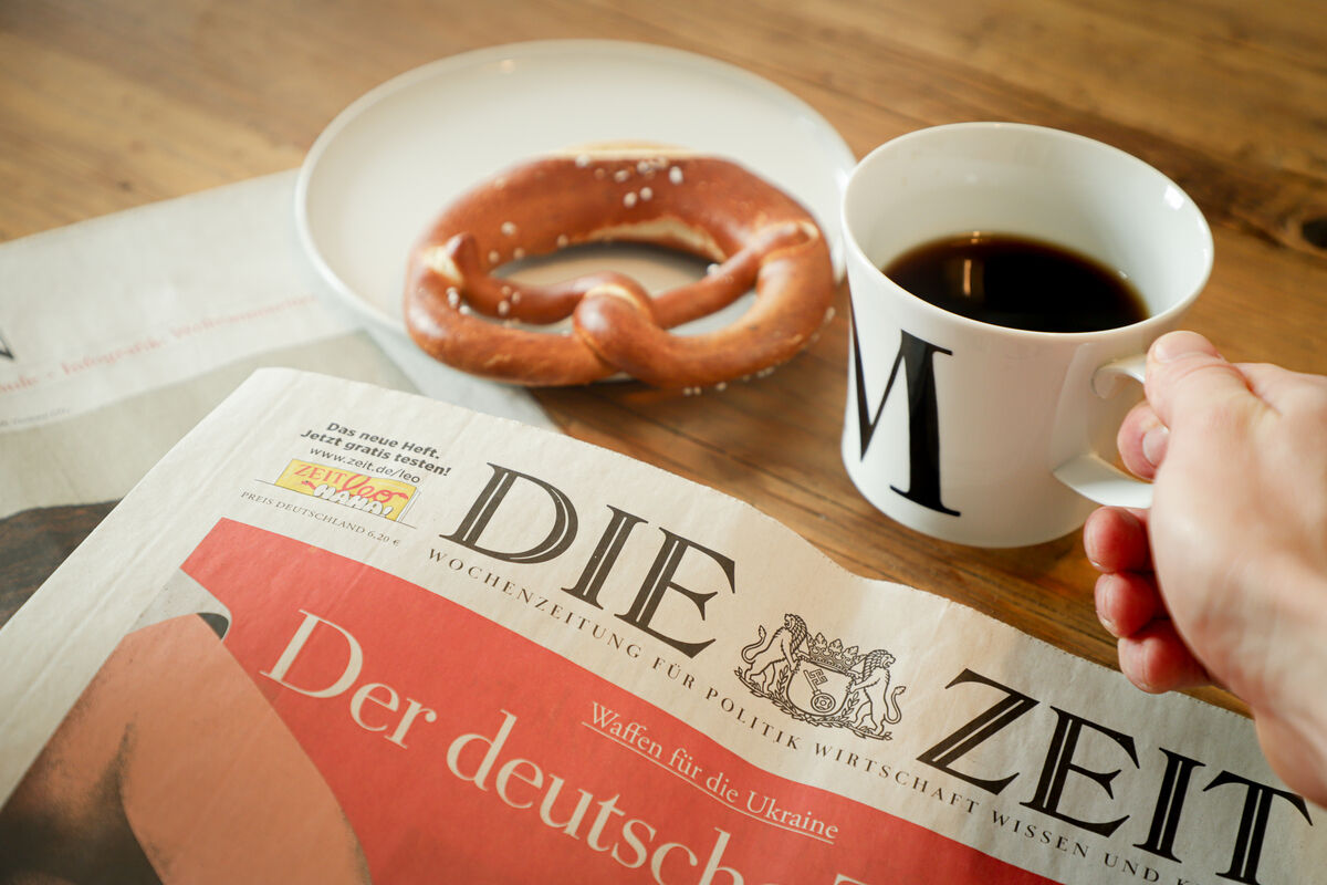 Eine weiße Frühstückstasse mit dem Buchstaben 'M' darauf, gefüllt mit Kaffee. Daneben liegt eine Brezel auf einem weißen Teller und eine Zeitung mit dem Titel 'DIE ZEIT' auf dem Tisch. Eine Hand hält die Tasse. Das Bild vermittelt eine gemütliche Morgenstimmung beim Frühstücklesen.