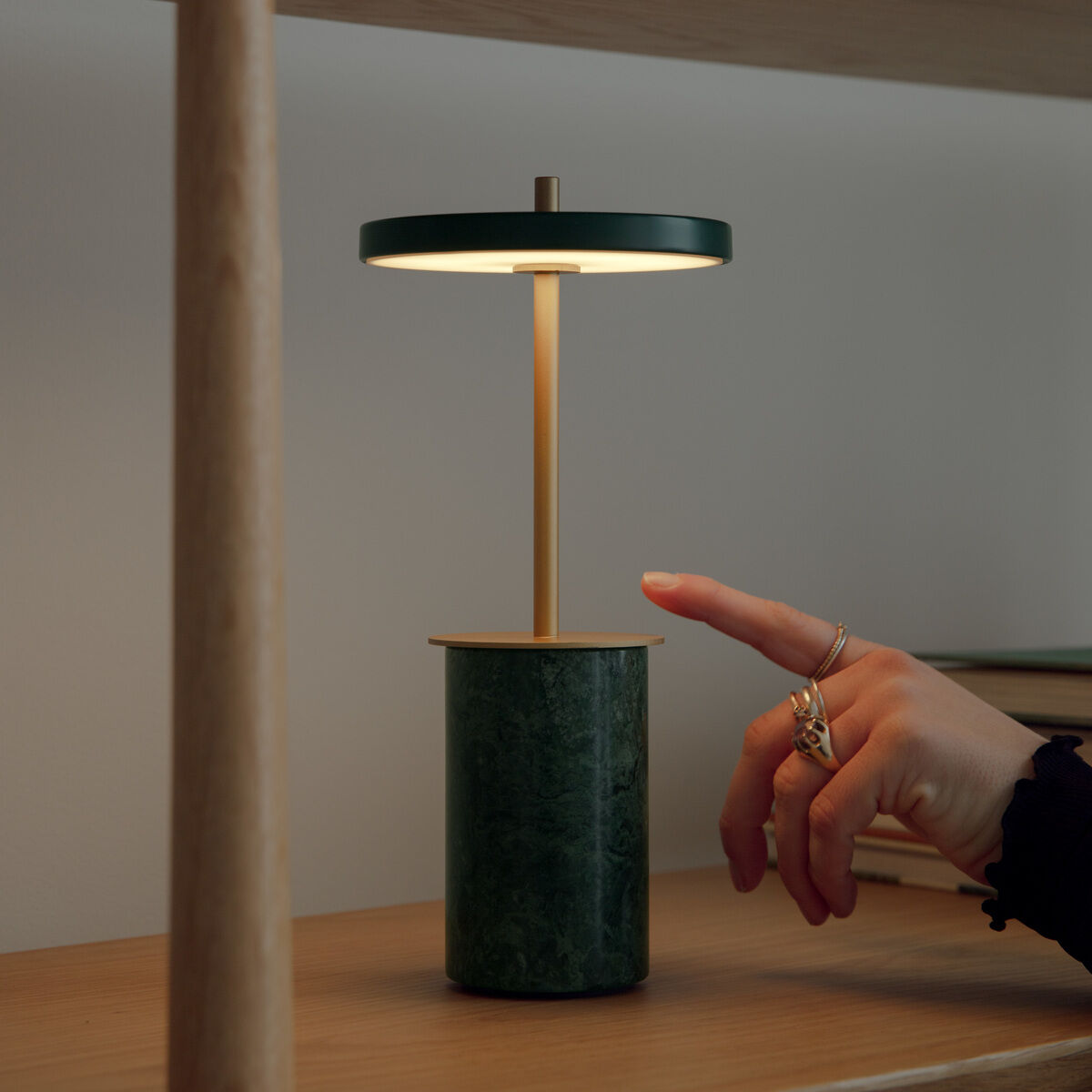 Kleine moderne grüne Tischlampe Asteria Move Mini steht auf einem holzfarbenen Regal. Die Lampe wird per Fingerdruck eingeschaltet. Im Hintergrund liegen Bücher. Das Bild vermittelt ein elegantes, minimalistisches Wohnambiente.