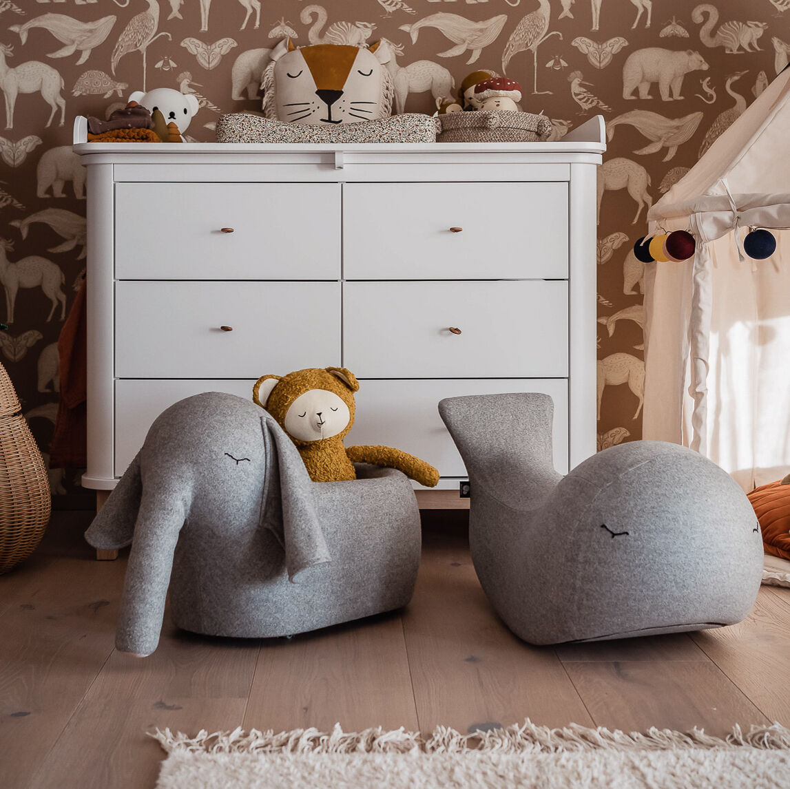Ein gemütlich eingerichtetes Kinderzimmer mit einem weißen Wickeltisch, dekoriert mit Kuscheltieren, und zwei weichen Rolltieren in Form eines Elefanten und eines Wals. Die Wände sind mit hellbrauner Tapete mit weißen Tiermotiven bedeckt.