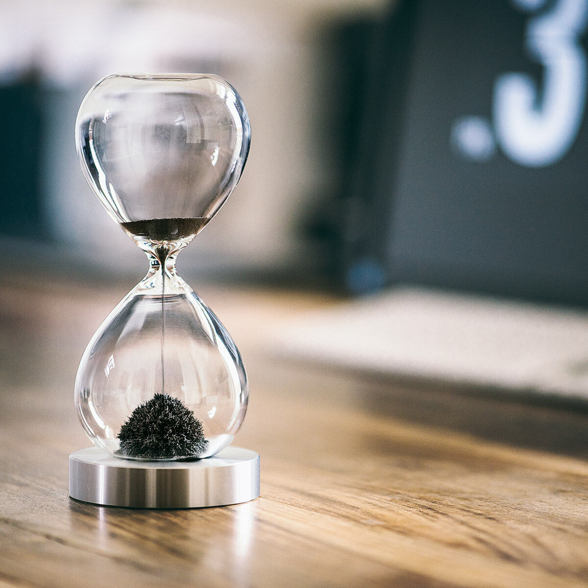 Eine magnetische Sanduhr mit schwarzem Sand auf einem Chromständer, platziert auf einem Holztisch in einem modernen Raum.