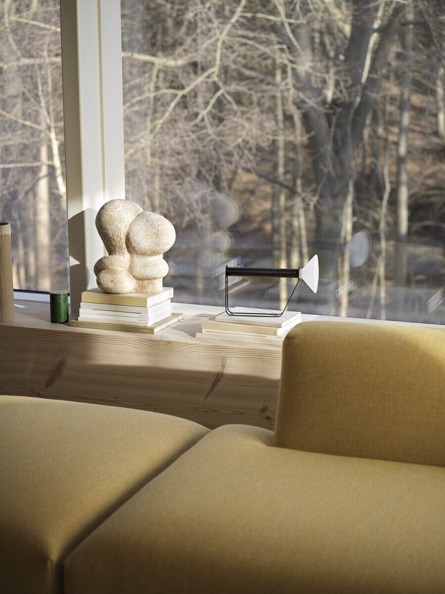 Ein modernes Wohnzimmer mit einer Tragbaren Lampe 'Piton' auf einer Holzbank neben einer kunstvollen Skulptur und mehreren Büchern vor einem großen Fenster mit Blick auf einen Wald.