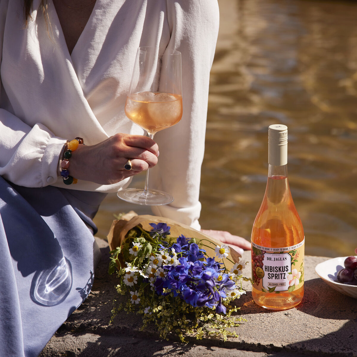 Drei Personen genießen einen entspannten Moment am Wasser mit einer Flasche Hibiskus Spritz, Blumen und Trauben. Eine Person hält ein Weinglas mit roséfarbenem Getränk, während eine andere nach Trauben greift.