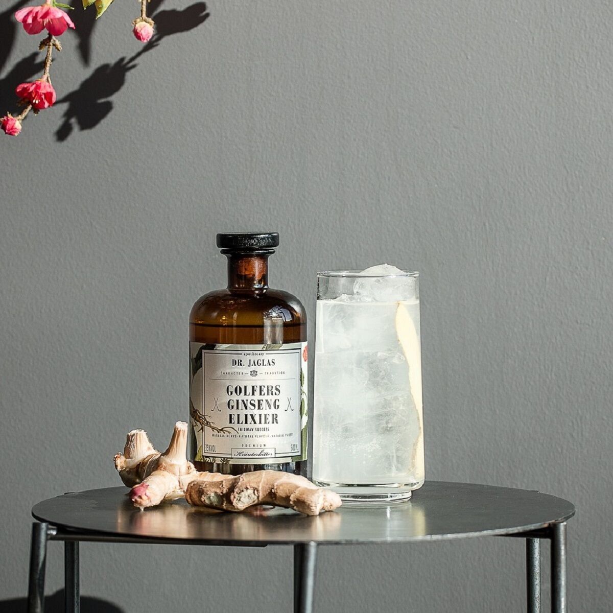 Eine Flasche Golfers Ginseng-Elixier von Dr. Jaglas, umgeben von frischem Ginseng, steht neben einem Glas gefüllt mit Eiswürfeln auf einem grauen Tisch, vor einem dunkelgrauen Hintergrund.