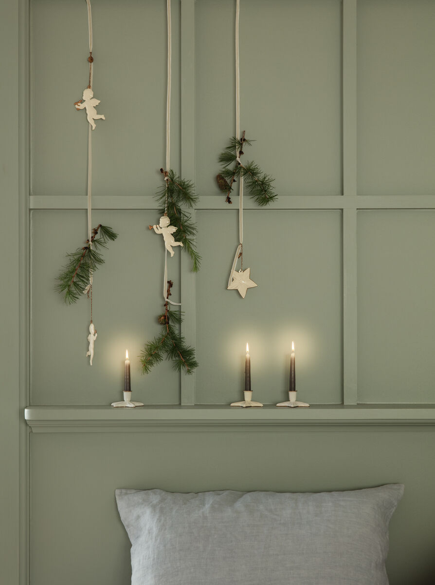 Ein Set von Weihnachtsanhängern namens 'Heilige Nacht', dekoriert mit Engeln und Sternen aus naturfarbenem Material, hängt an einer grünen Wand, geschmückt mit Tannenzweigen. Darunter stehen drei brennende Kerzen in schlichten Haltern.