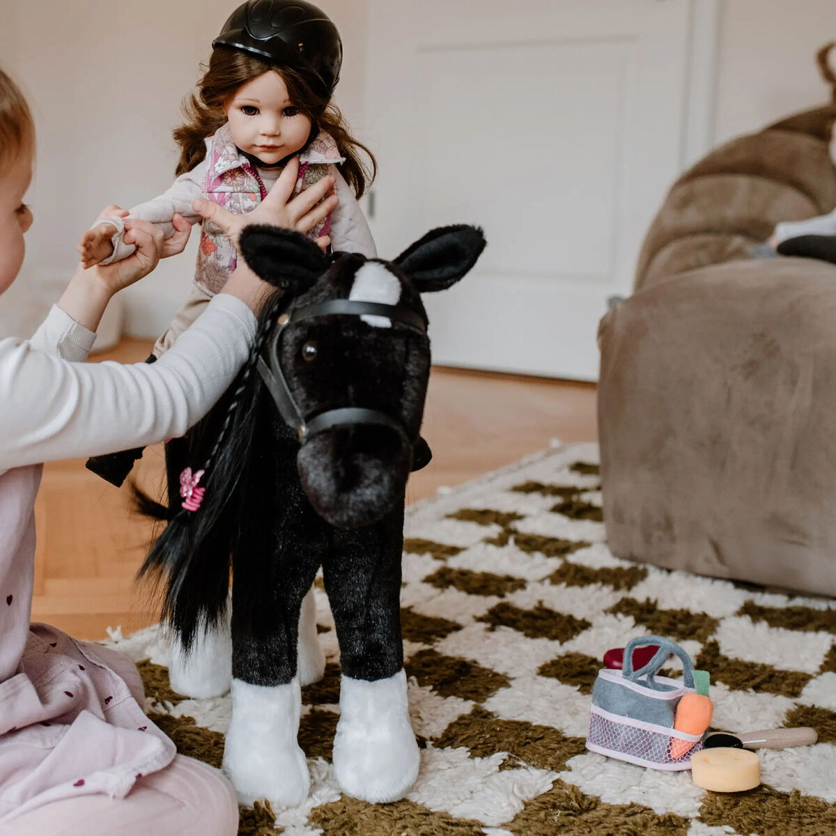 Ein Kind spielt mit der Stehpuppe »Hannah Loves Horseback Riding« von Götz Puppenmanufaktur, die Reitkleidung trägt und auf einem großen schwarzen Stoffpferd sitzt. Im Hintergrund: Teppich, beigefarbenes Sofa, Spielzeugkorb. Warme, einladende Atmosphäre.