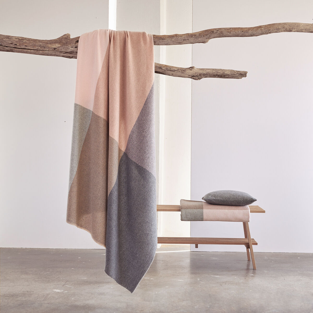Auf einem rustikalen Ast hängt eine elegante Merino-Kaschmirdecke »Monte Carlo« von Eagle Products in gedeckten Beige-, Rosa- und Grautönen. Im Hintergrund steht eine Holzbank, darauf ein passendes Kissen und eine weitere gefaltete Decke, minimalistisches Ambiente.