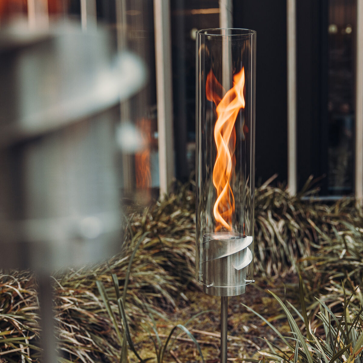Ein moderner Erdspieß mit einem Glaszylinder, der eine brennende Flamme enthält, ist in einem Garten mit grünen Pflanzen und hohem Gras zu sehen. Der Hintergrund zeigt eine moderne Gebäudestruktur mit vertikalen Fenstern.