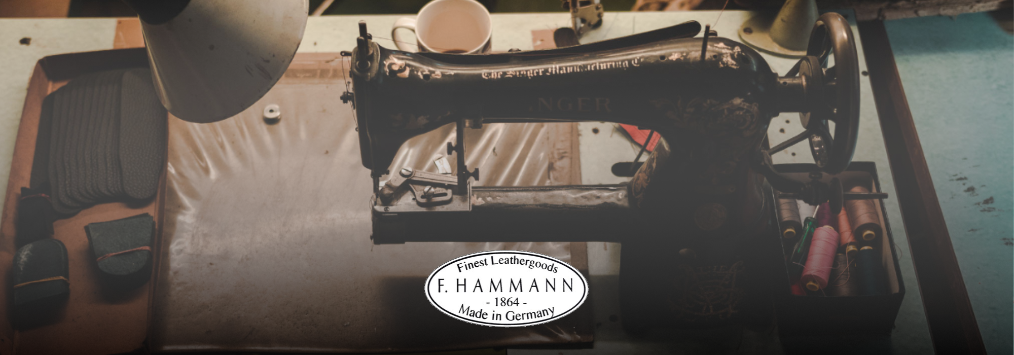 Alte schwarze Nähmaschine auf einem Arbeitstisch mit einer Tasse im Hintergrund und einem Logo 'F. Hammann seit 1864 Made in Germany'