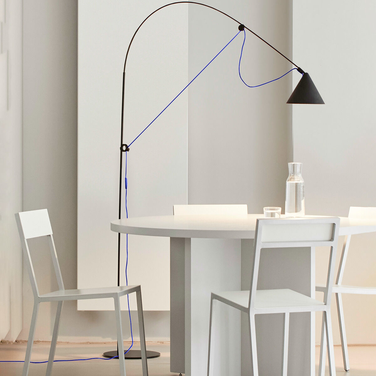 Eine moderne Stehlampe in Schwarz mit einem ultramarinblauen Kabel, die neben einem weißen Tisch und Stühlen steht. Das Design ist minimalistisch und elegant, ideal für moderne Wohn- oder Arbeitsräume.