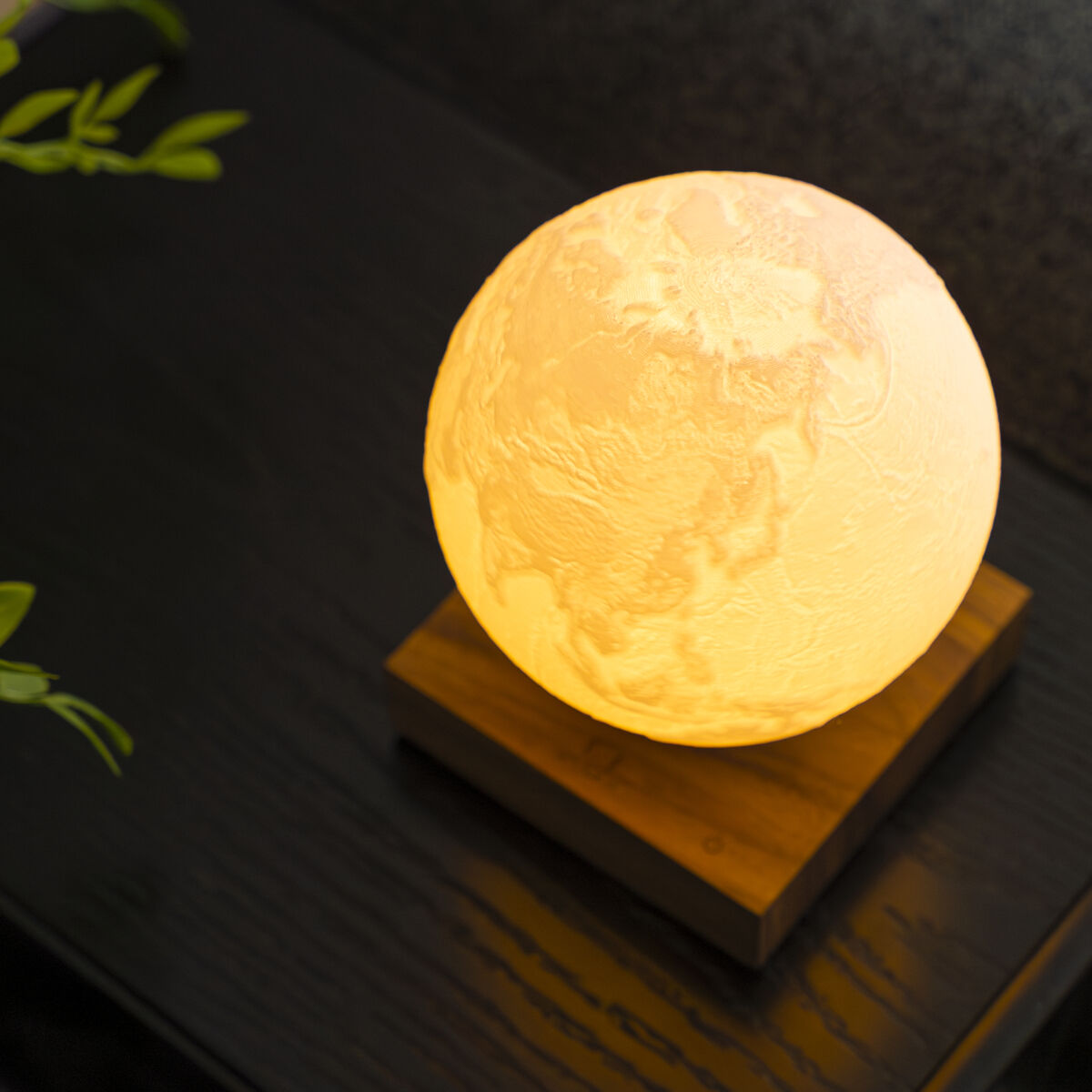 Die Leuchte »Smart Earth Lamp« von Gingko Design zeigt eine detaillierte leuchtende Weltkugel auf einem schlichten rechteckigen Holzsockel. Das sanfte, warme Licht erzeugt eine gemütliche Atmosphäre. Sie steht auf einem dunklen Holztisch, links sind grüne Blätter zu sehen.