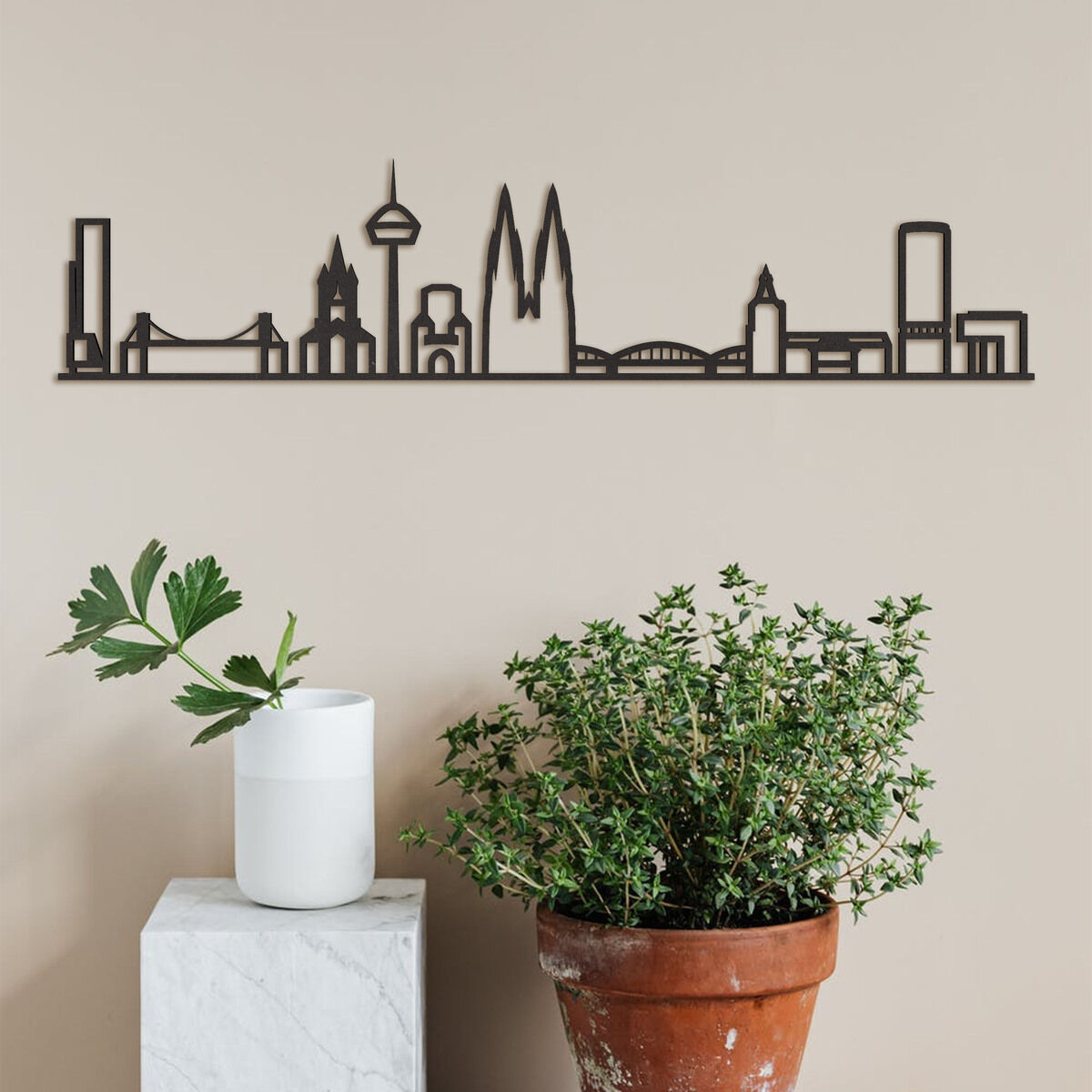 Holz-Skyline von Köln, schwarze Silhouette von Wahrzeichen wie dem Kölner Dom und dem Fernsehturm, an einer beige Wand hängend. Unten links ein weißer Blumentopf mit einer Pflanze und rechts ein größerer Terrakotta-Topf mit grüner Flora.