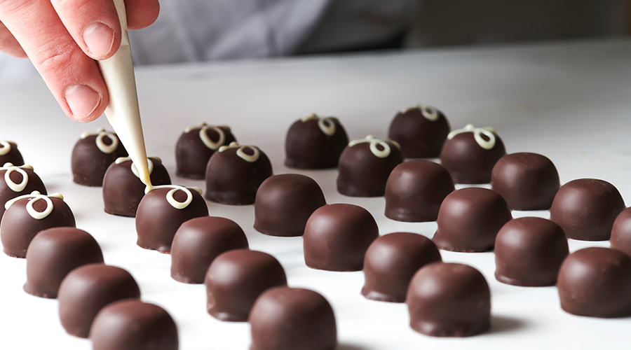 Hand verziert dunkle Schokoladenpralinen mit weißer Schokoladendekoration auf weißer Oberfläche