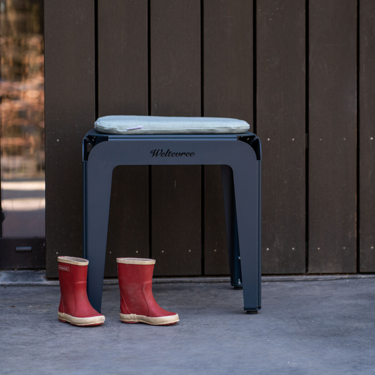 Ein moderner Hocker namens »Bended Stool«, platziert vor einer Wand mit Holzoptik. Neben dem Hocker stehen rote Gummistiefel auf einem grauen Boden. Das Gesamtdesign wirkt minimalistisch und funktional.