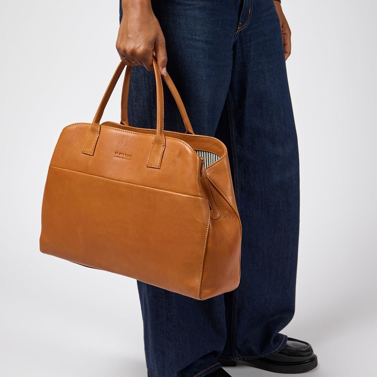 Eine hellbraune Schultertasche aus Narbenleder der Marke O My Bag wird von einer Person an der Hand getragen. Die Tasche hat zwei stabile Henkel, ein schlichtes Design und ein gestreiftes Innenfutter. Die Person trägt eine dunkelblaue Hose und schwarze Schuhe.