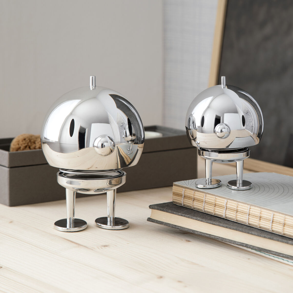 Zwei Hoptimist Bumble Chrome Figuren stehen auf einem hellen Holztisch. Eine Figur ist größer als die andere, beide haben ein glänzendes Chrom-Finish. Im Hintergrund befinden sich Bücher und Schachteln in neutralen Farben, was der Aufnahme ein modernes und minimalistisches Flair gibt.