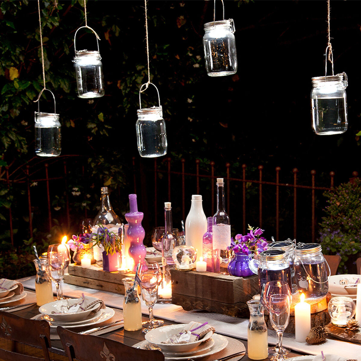 Ein festlich gedeckter Tisch im Freien bei Nacht, dekoriert mit hängenden Sonnenglas Classic Solarleuchten und Kerzen. Der Tisch ist mit Geschirrsets, lila und weißen Flaschen sowie verschiedenen Dekorationen geschmückt. Eine gemütliche und einladende Atmosphäre für eine Gartenparty.