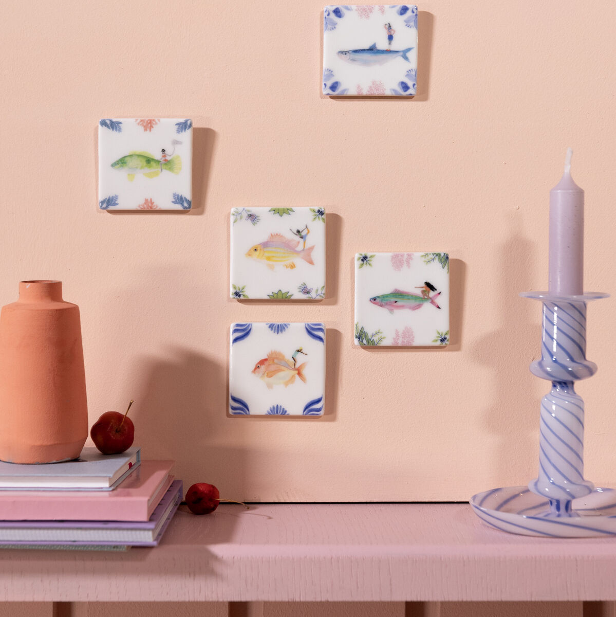 Eine Sammlung von fünf dekorativen Keramikfliesen mit Meeresmotiven an einer beigen Wand. Die Fliesen zeigen Bilder von Meerestieren und sind mit blauen und grünen Details verziert. Eine orange Vase, gestapelte Bücher und eine blau-weiße gestreifte Kerze stehen auf einem Regal darunter.
