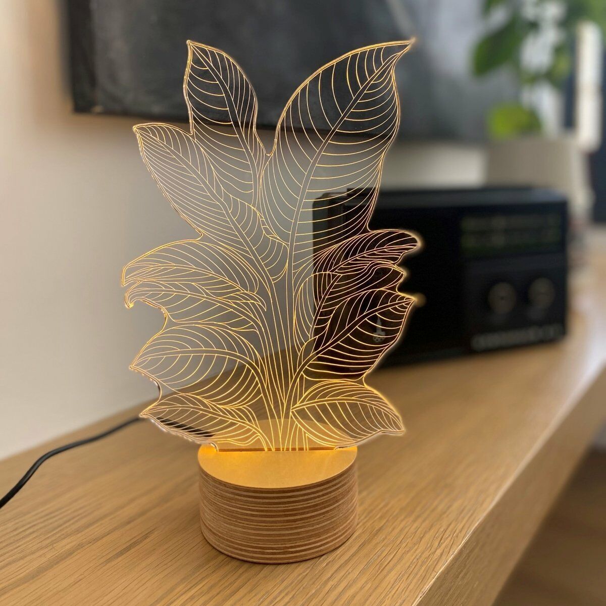 3D-Lampe im Heliconia-Design mit einer geschnittenen Holzbasis, die eine filigrane, goldene Pflanzenform darstellt und auf einem Tisch in einem Wohnzimmer steht.
