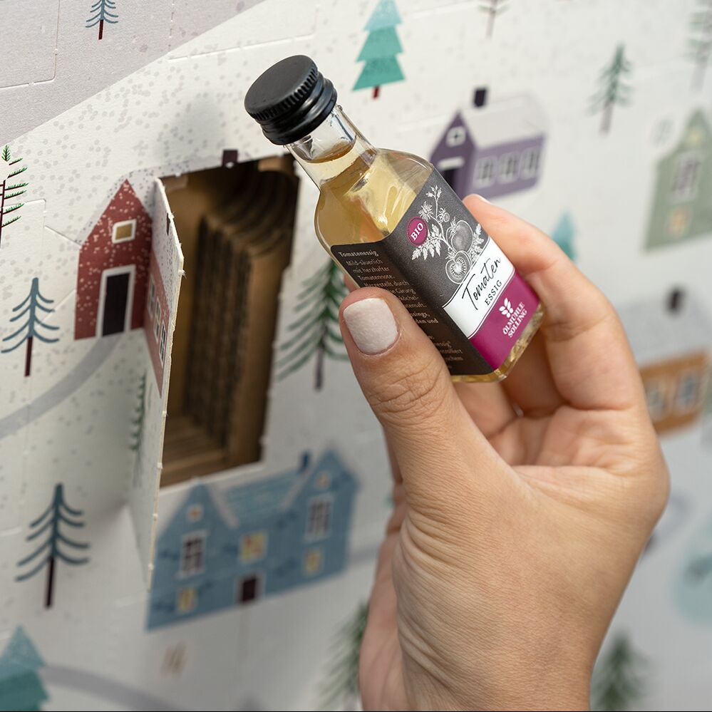 Nahaufnahme eines Adventskalenders mit winterlicher Dorf- und Schneelandschaft. Eine Hand öffnet ein Türchen und hält eine kleine Flasche in der Hand. Der Kalender ist dekorativ mit Häusern und Tannenbäumen gestaltet.