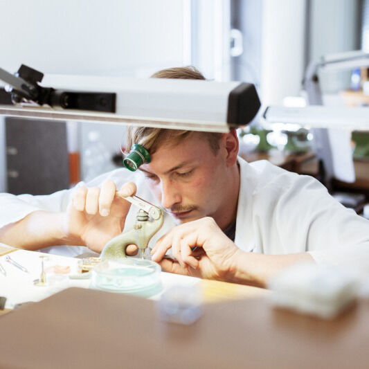 ZEIT-Sonderedition »Flieger Automatic Bronze« Uhr mit einem Handwerker, der präzise an einem Uhrwerk arbeitet. Die Umgebung deutet auf eine Werkstatt oder ein Studio hin, mit verschiedenen Werkzeugen und Materialien im Hintergrund.