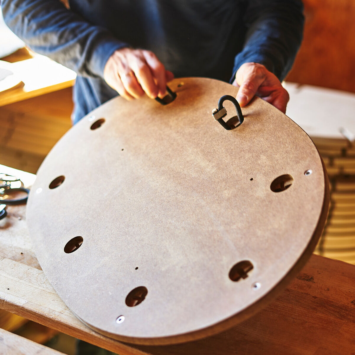 Eine Person hält ein rundes Sensoboard aus Sperrholz mit einem Cutout-Design. Das Balancebrett hat mehrere Löcher und zwei Griffe. Die Person trägt einen dunkelblauen Pullover. Im Hintergrund ist eine Holzstruktur zu erkennen.