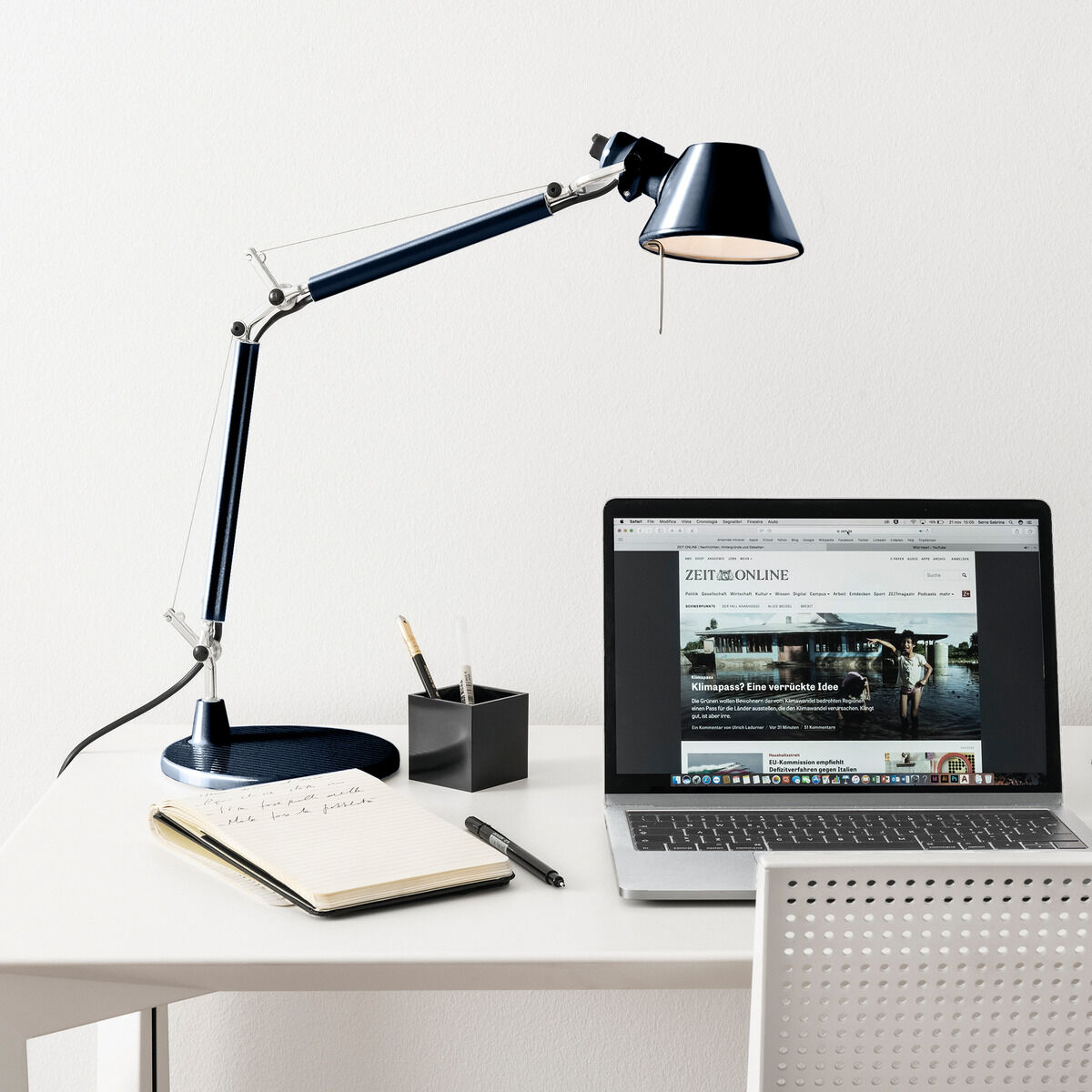 Eine elegante schwarze Tischlampe der ZEIT-Sonderedition »Tolomeo Micro« steht auf einem weißen Schreibtisch neben einem Laptop und einem Notizbuch. Der moderne Arbeitsplatz ist stilvoll eingerichtet und die verstellbare Lampe beleuchtet den Raum optimal.