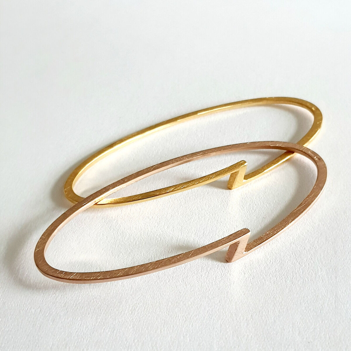 Zwei elegante Armreifen, einer in Gold und einer in Rosa, nebeneinander auf einer weißen Oberfläche.