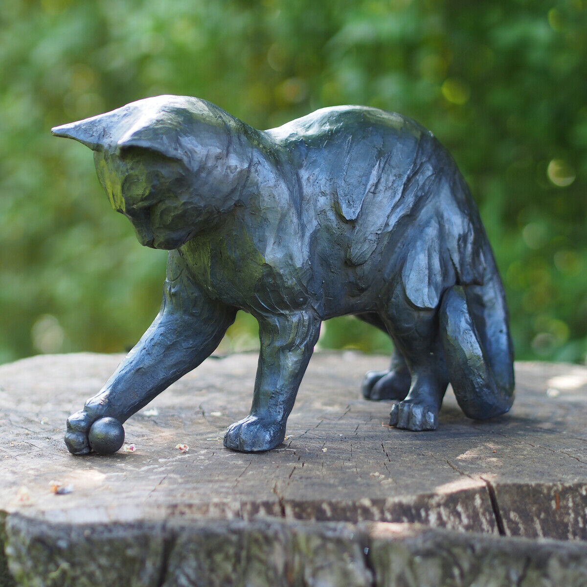 Bronzene Gartenskulptur einer verspielten Katze mit einem Ball auf einem Baumstumpf, umgeben von verschwommenem Grün.