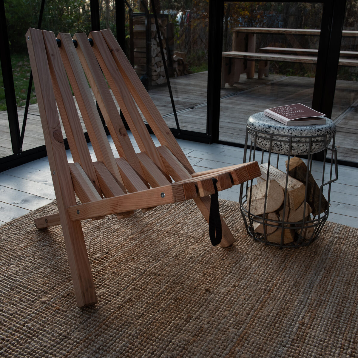 Ein Feldstuhl aus Holz mit Schafsfellüberzug steht auf einem gewebten Teppich in einem verglasten Raum mit Blick auf einen Garten.