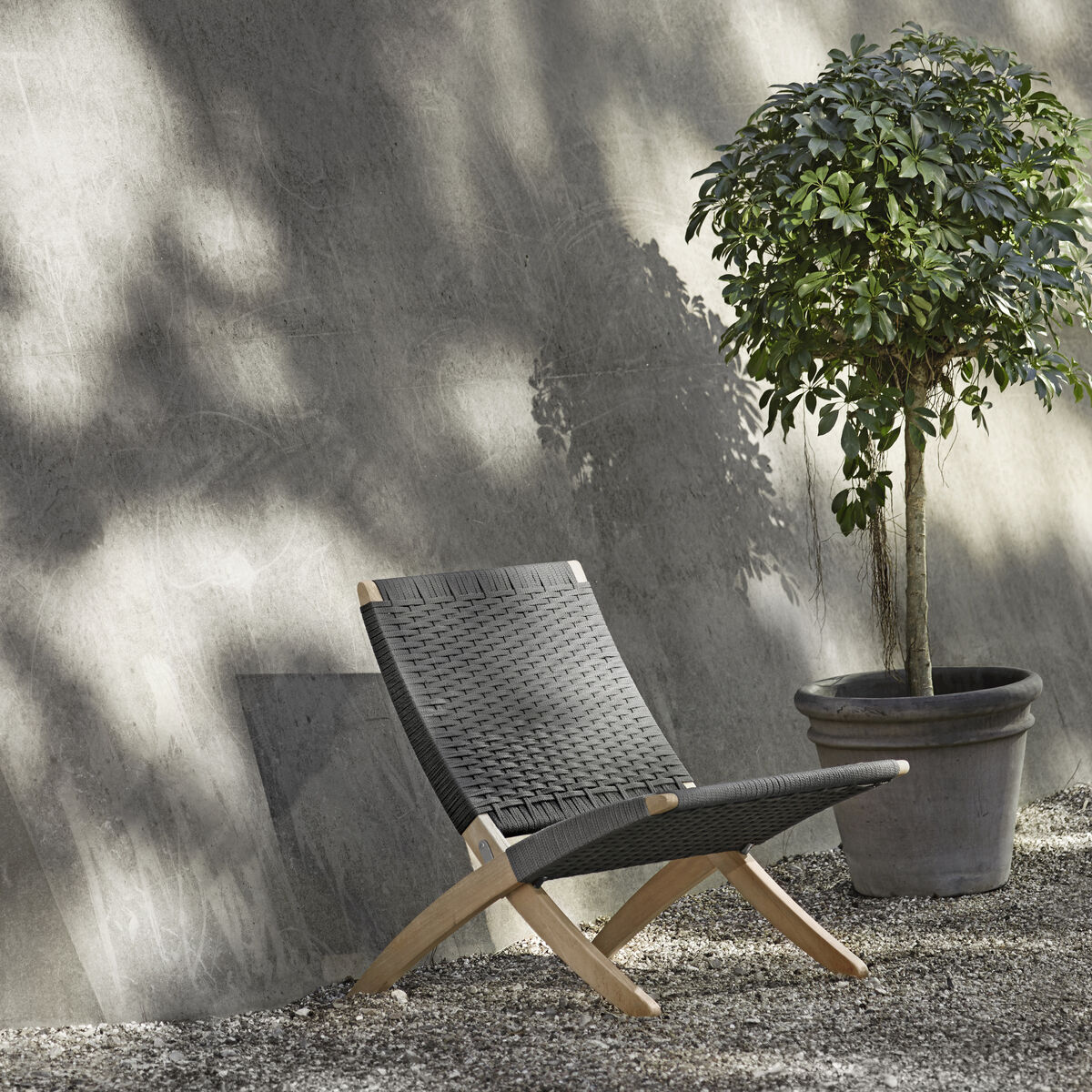 Ein moderner Outdoor-Stuhl mit schwarzem Sitz und braunem Holzrahmen steht neben einem grünen Blattpflanze in einem Topf vor einer grauen Wand mit Schattenspielen.