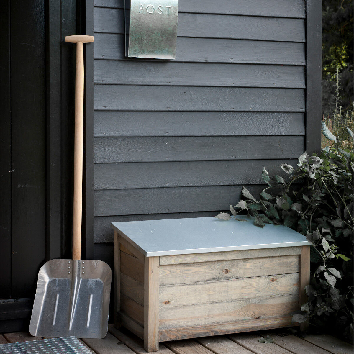 Bild einer rustikalen Holz-Gartenbox »Aldsworth« neben einer großen Metallschaufel und einem silbernen Briefkasten an einer grau gestrichenen Wand. Die Kiste hat einen Deckel in helleren Farben. Neben der Holzbox sind grüne Pflanzen zu sehen.