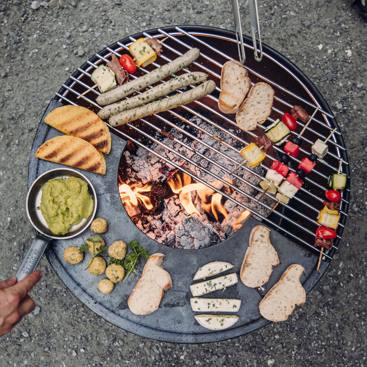 Eine Feuerschale mit Grillrost voller verschiedener Lebensmittel, darunter Brot, Würstchen und Gemüse-Spieße, wird von einer Hand mit einer Pfanne mit Essen daneben gehalten. Rundherum stehen mehrere Personen auf einem Schotterboden.