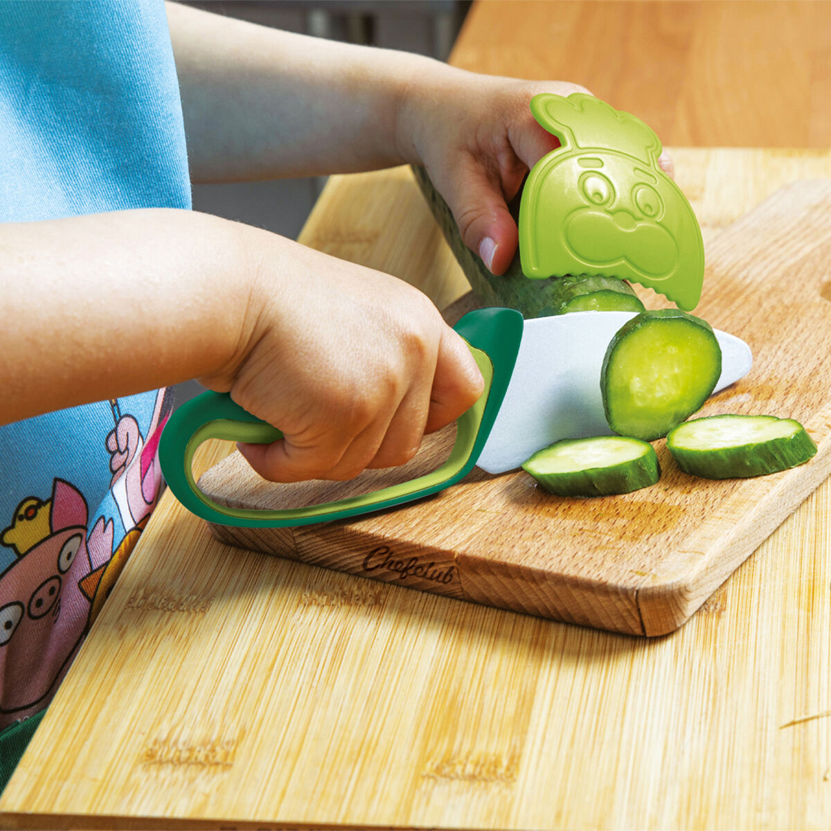 Ein Kind benutzt ein grünes Kindermesser mit einem fröhlich aussehenden Schutzaufsatz, um eine Gurke auf einem Holzbrett zu schneiden. Der Hintergrund zeigt eine bunte Schürze und eine Küche.