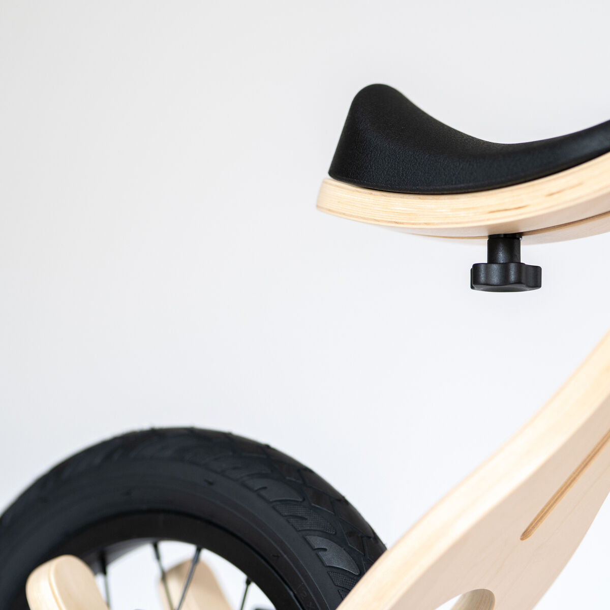 Detailaufnahme eines 3in1 Balance Bikes für Kinder aus hellem Holz mit schwarzem Sattel und einem Teil des Hinterrads, minimalistisches und modernes Design auf hellem Hintergrund.