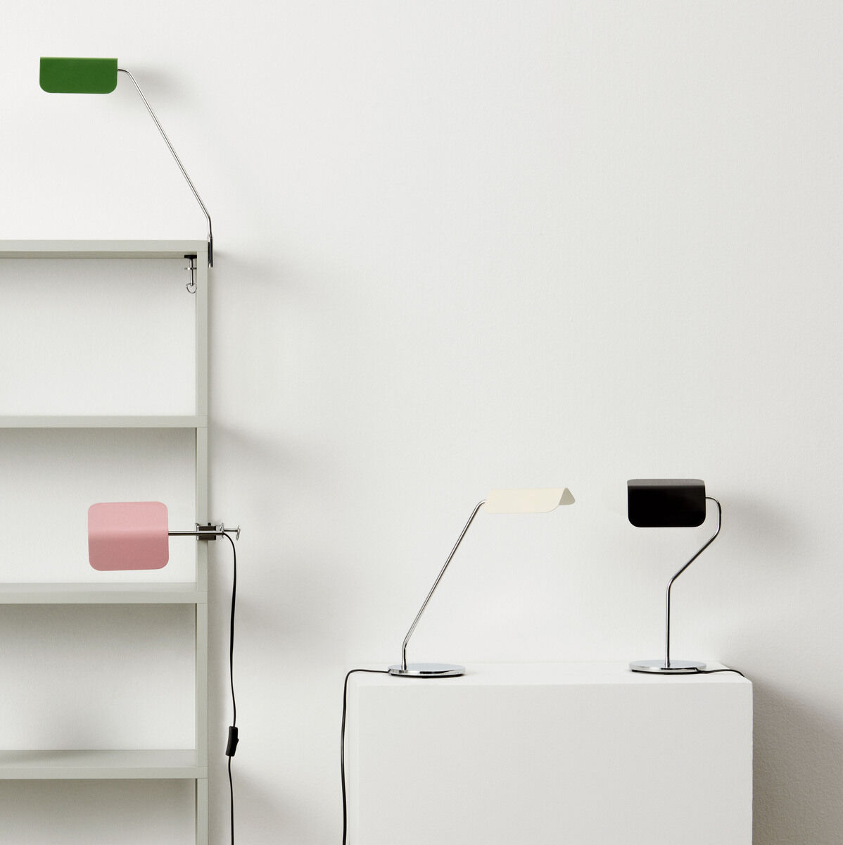 Mehrere moderne Schreibtischlampen, jede in einer anderen Farbe (Schwarz, Grün, Rot, Blau und Rosa), sind auf einem Regal und einem weißen Podest präsentiert. Die Lampen haben einen minimalistischen Stil mit verstellbaren Armen.