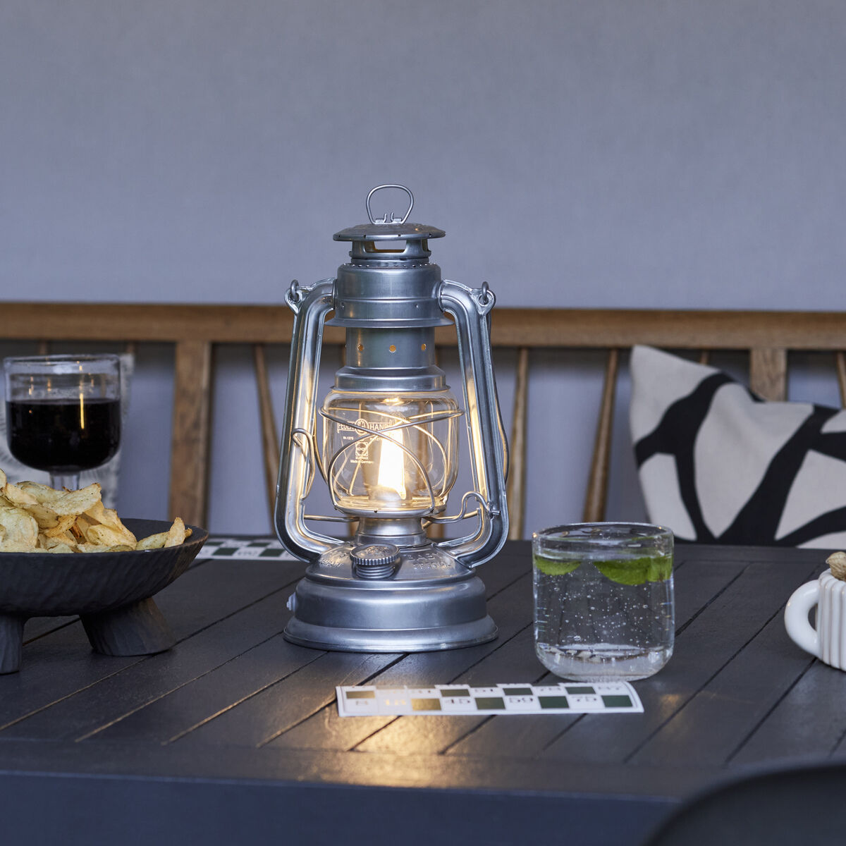 Eine metallische LED Laterne namens Baby Special 276 steht auf einem dunklen Holztisch. Die Laterne ist im Vintage-Stil gehalten und umgeben von Snacks und Getränken. Im Hintergrund ist eine Bank mit Kissen zu sehen.