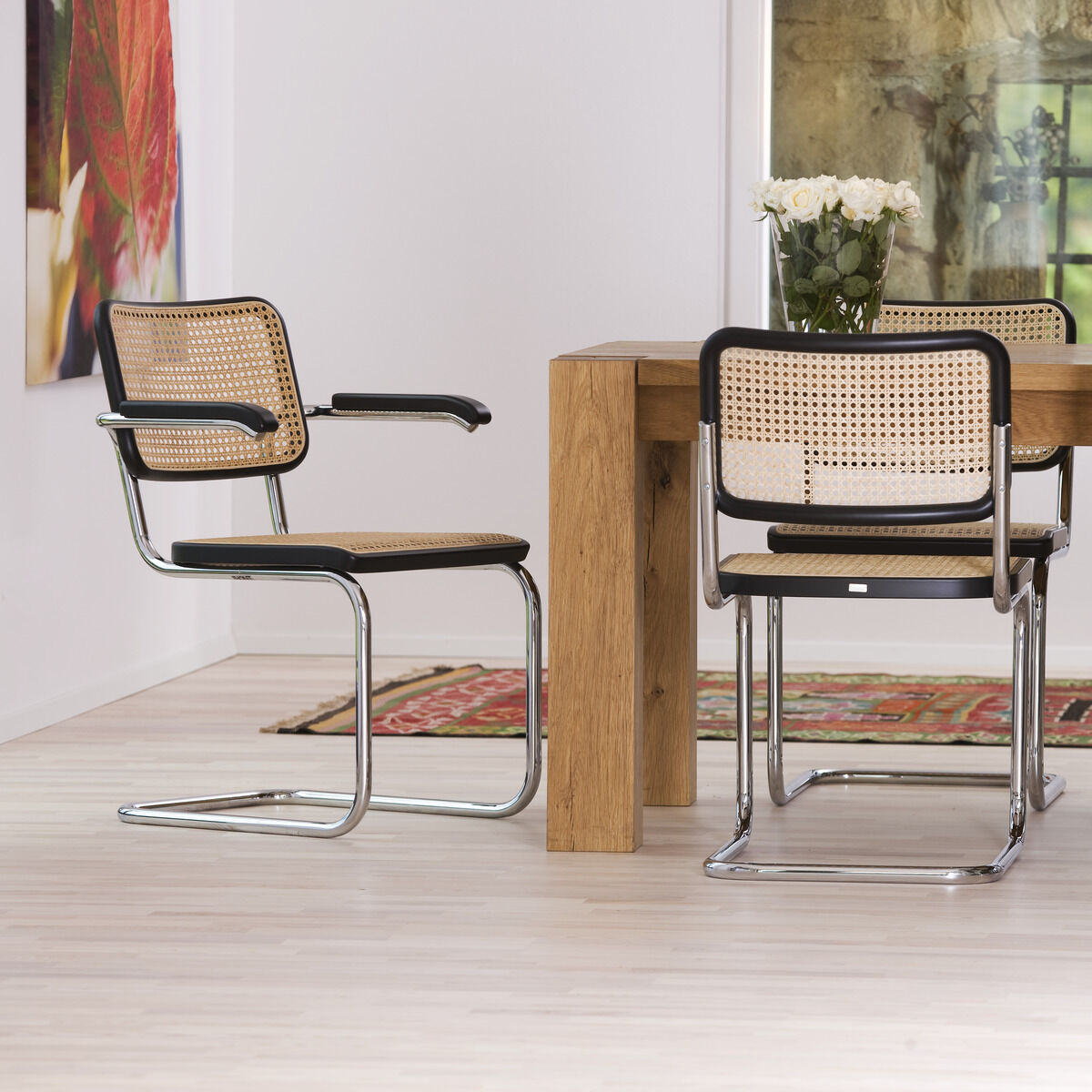 Esszimmer mit Holztisch und vier Freischwinger-Stühlen aus Stahlrohr und Rattan der Marke Thonet. Moderne und minimalistische Einrichtung mit hellen Wänden und Kunstwerken im Hintergrund.