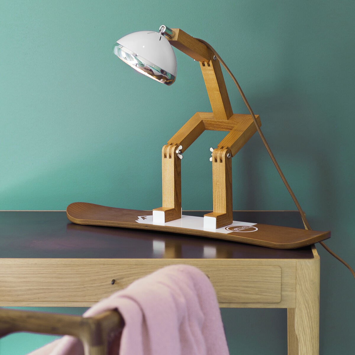 Eine Designer-Tischlampe in Form eines kleinen Holzmännchens sitzt auf einem Skateboard, das als Sockel dient. Die Lampe hat ein minimalistisches, moderne Design und steht auf einem Holztisch. Im Hintergrund ist eine grüne Wand zu sehen und über einem Stuhl hängt eine rosafarbene Decke.