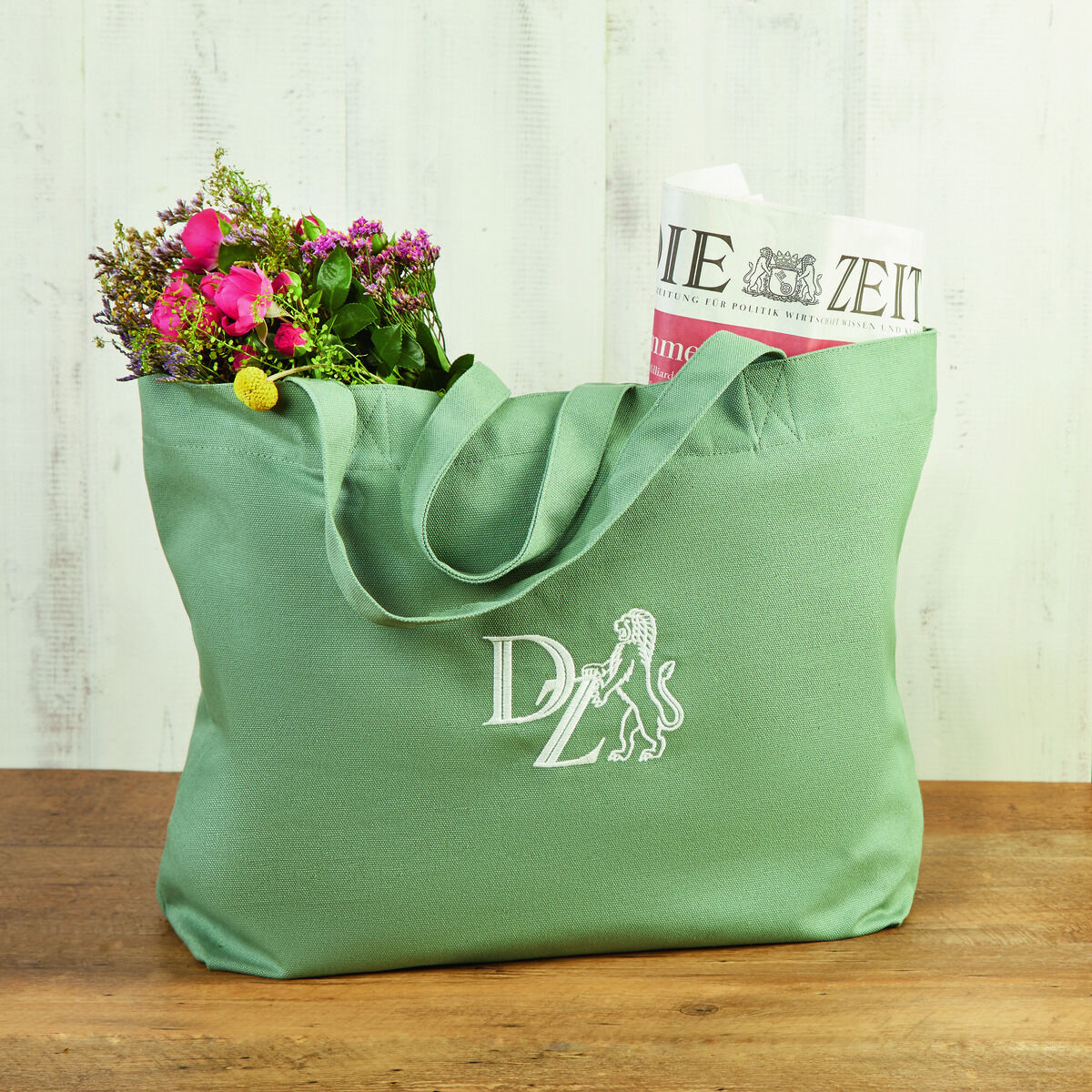 Grüne Canvas-Tasche mit Weißdruck, die Blumen und eine Zeitung enthält, auf einem Holztisch vor einer hellen Holzwand.