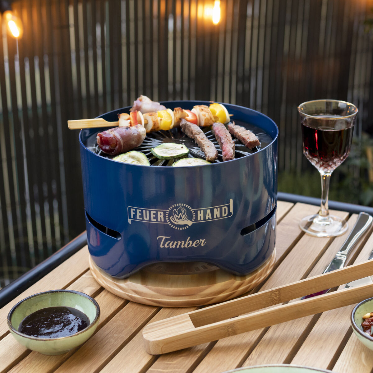 Tischgrill 'Tamber' von Feuerhand auf einem Holztisch im Freien, dekorativ dekoriert. Surrounding Grillbesteck, ein Glas Rotwein und gegrilltes Gemüse auf Tellern. Balkondekorationen und beleuchtete Lichter im Hintergrund.