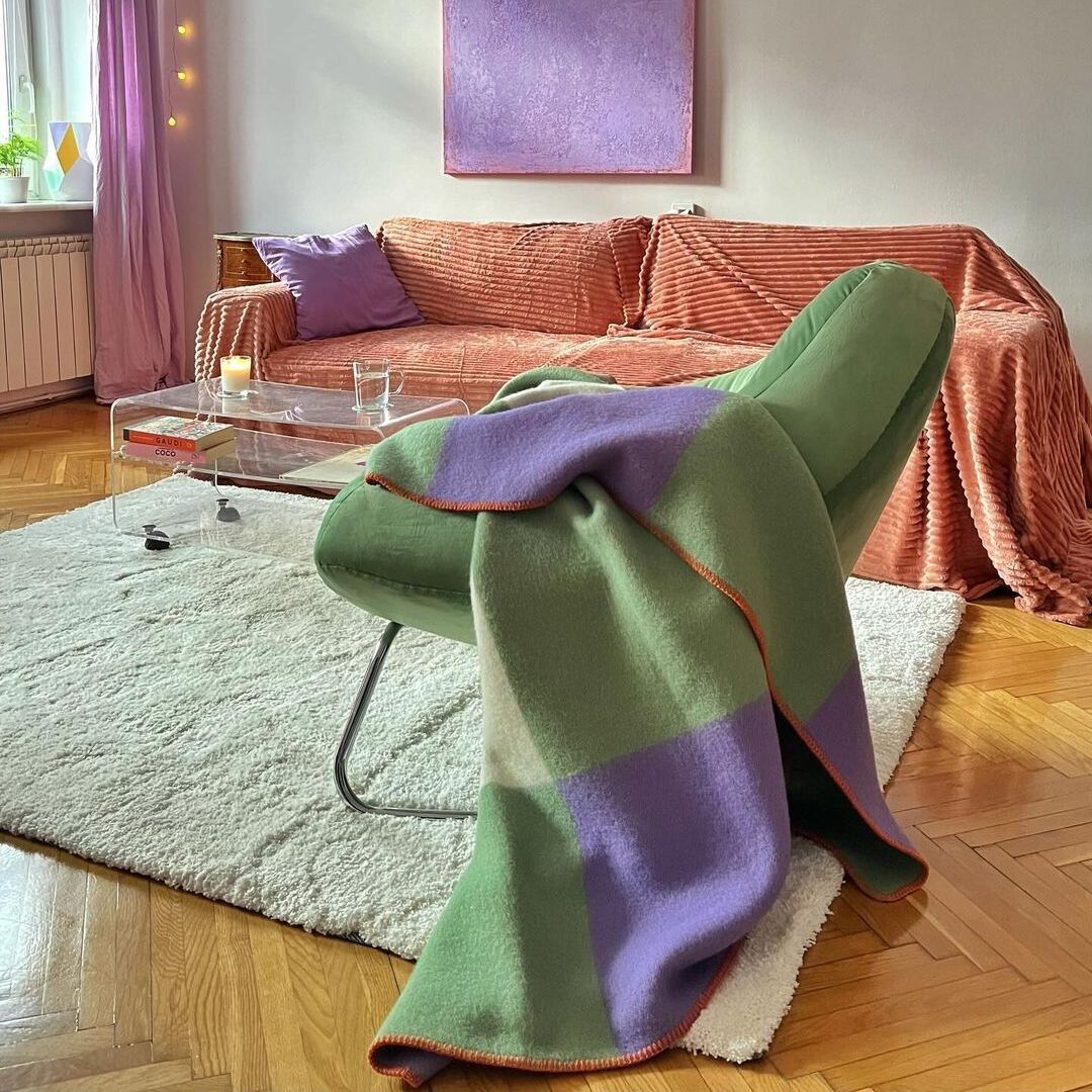 Ein gemütliches Wohnzimmer mit einem grünen, lila und orangefarbenen Plaid über einem modernen Stuhl. Ein rosa Sofa, ein Glastisch und eine Lichterkette an der Wand sorgen für eine entspannte Atmosphäre.