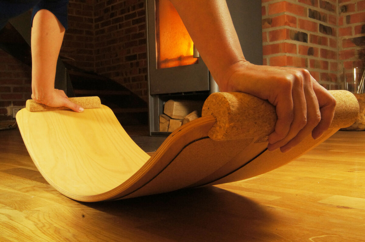 Hände, die ein Balance Board aus Holz und Kork auf einem Holzboden halten, im Hintergrund ein Kamin und Ziegelwände.