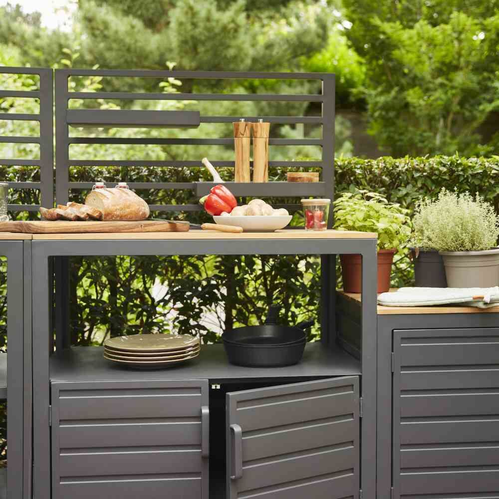 Outdoor-Küchen-Mittelteil 'Sierra' im Garten mit grüner Hecke, grauem Design und Holzplatte; Geschirr und Kochutensilien vorhanden.