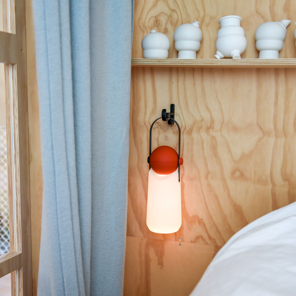 Eine tragbare Leuchte namens "Guidelight" der Marke Weltevree hängt an einer hellen Holzvertäfelung neben hellblauen Vorhängen und einem weißen Bett. Die Leuchte hat einen orangefarbenen oberen Teil, einen matten, zylindrischen Leuchtenkörper und einen schwarzen Henkel. Darüber stehen weiße Keramikgefäße auf einem schmalen Regal.