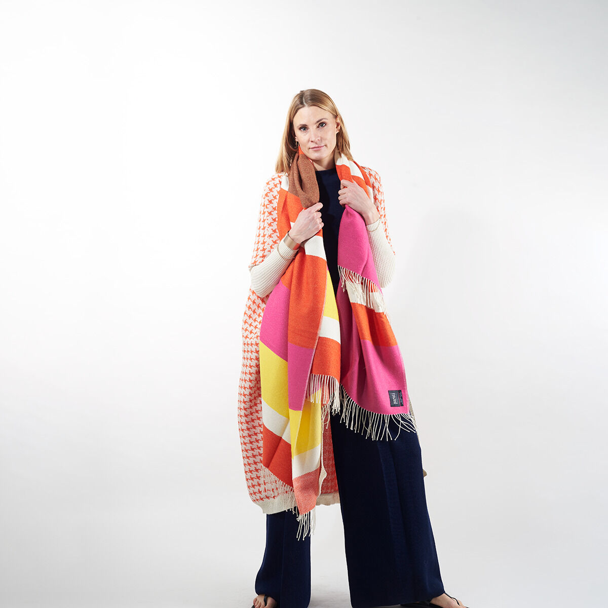 Eine Frau trägt eine ZEIT-Jubiläumsedition Cape mit dem Namen »Surya«. Das Cape ist farbenfroh mit Pink-, Orange- und Weißtönen gestaltet. Die Frau steht vor einem neutralen Hintergrund und trägt einen dunklen, langen Rock oder Mantel.