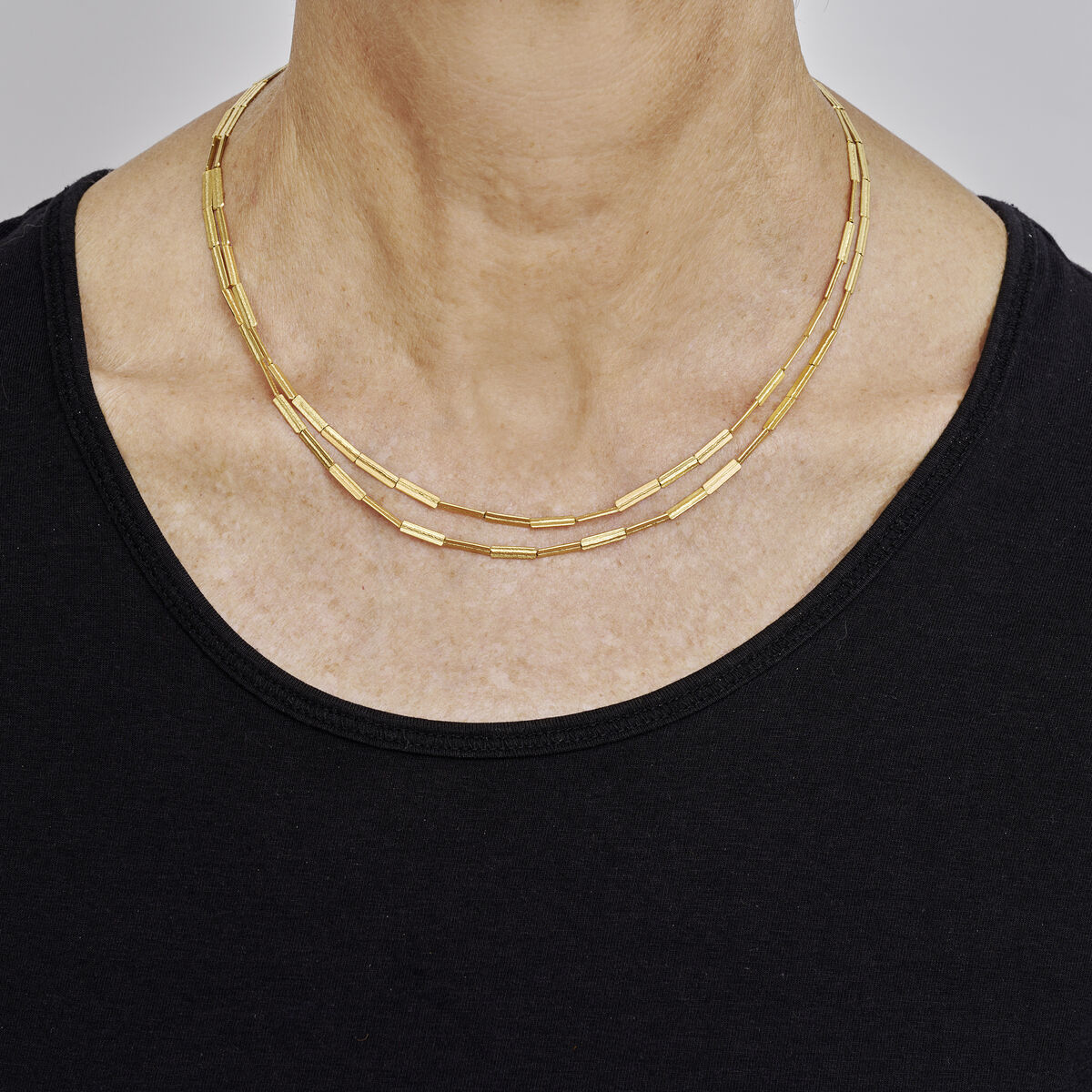 Eine fein goldplattierte, silberne Halmkette mit geprägten, rechteckigen Gliedern wird um den Hals einer Person getragen. Das Collier liegt in zwei Reihen übereinander an und hebt sich elegant von dem schwarzen Oberteil der Person ab. Der Hintergrund ist neutral grau.