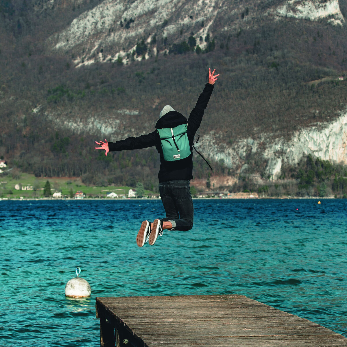 Person mit grüner Fahrradtasche »Annecy« springt von einem Holzsteg ins Wasser, im Hintergrund ein See und Berge.