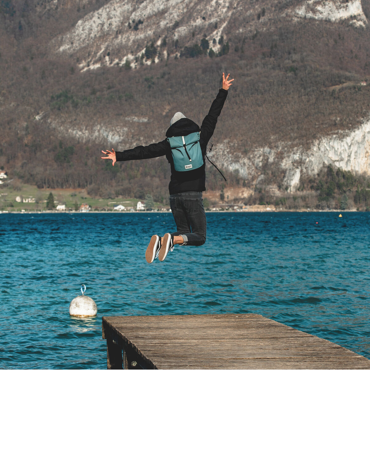 Person mit grüner Fahrradtasche »Annecy« springt von einem Holzsteg ins Wasser, im Hintergrund ein See und Berge.