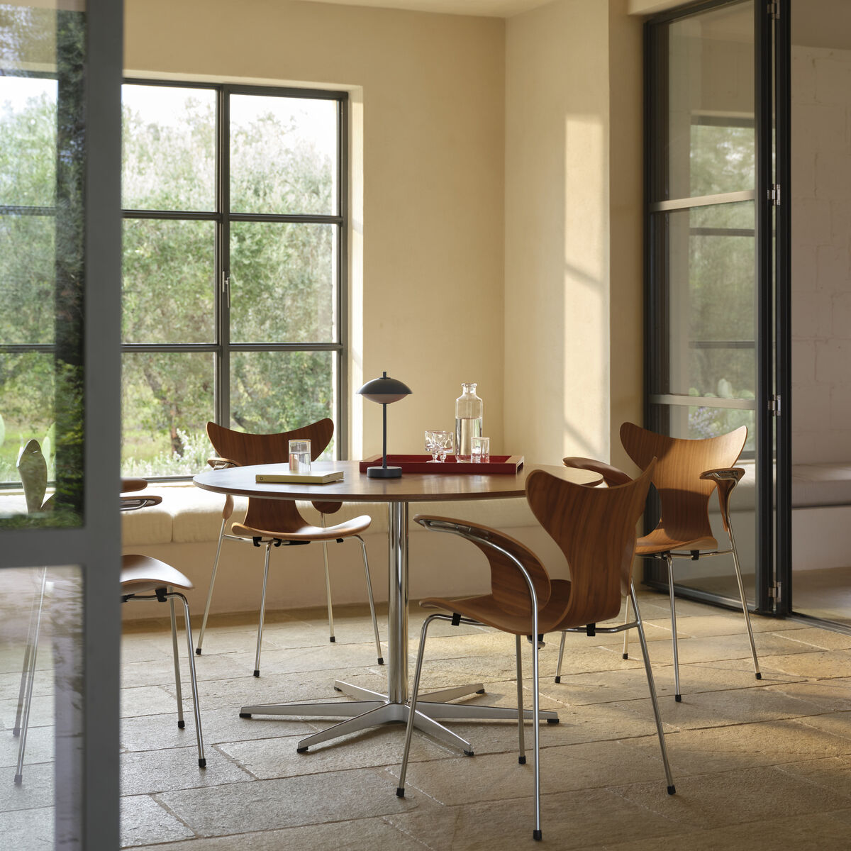 Heller Raum mit großen Fenstern und sandfarbenen Wänden. Im Mittelpunkt steht ein runder Tisch aus Holz und Metall mit vier eleganten Holzstühlen. Auf dem Tisch stehen die blaue tragbare Lampe »Clam™« von Fritz Hansen, Gläser und Karaffen. Hinter dem Tisch sieht man Grünpflanzen draußen.