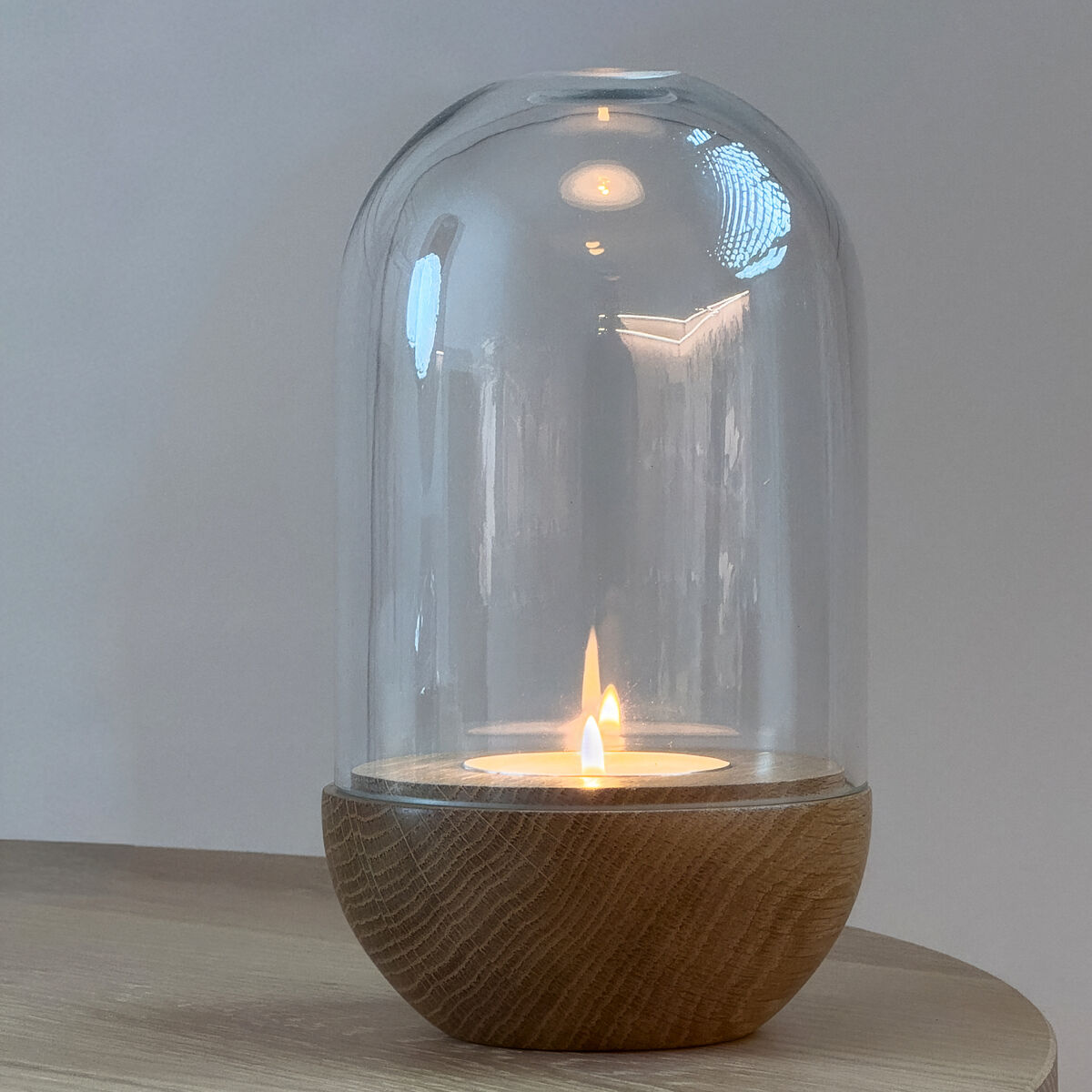 Windlicht »GLOW« von finbeaux, Variante Klarglas: Mundgeblasenes, transparentes Glas mit massivem Eichenholzfuß. In der Mitte brennt eine Kerze, die Licht wirft. Steht auf rundem Holztisch vor einfarbig hellem Hintergrund. Schlichtes, elegantes Design.